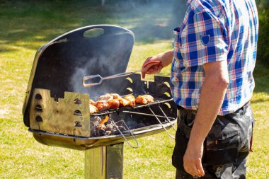 Kömür lokantasındaki bir adam ızgarada pişmiş çiğ eti ızgaraya koymak için ızgara maşa kullanıyor. Güneşli bir bahar gününde yeşil çimenli arka planda ızgaraya odaklan. Mangal konsepti. Makro.
