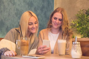 İki genç kız cep telefonuyla kafe ve içki cof gülümseyen