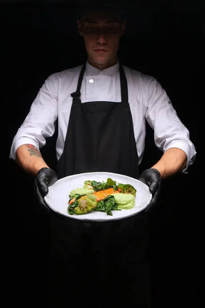 male cook in a black apron holds a plate with cooked bird fillet