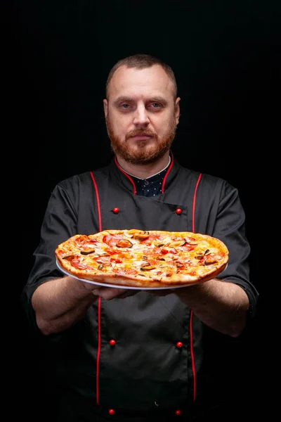 Siyah ceketli bir erkek aşçı ve eldivenler önünde zeytinli taze bir pizza tutuyor.