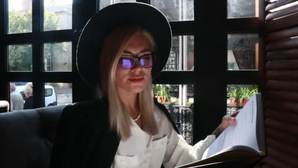 金发女人戴着黑色的帽子坐在餐馆的桌子旁 — 图库视频影像
