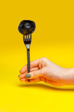 Siyah bir çatal üzerinde siyah bir hamur tatlısı, sarı bir arka planda kadın eli.