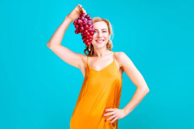 Stüdyoda mavi arka planda bir demet üzüm tutan sarışın kadın.