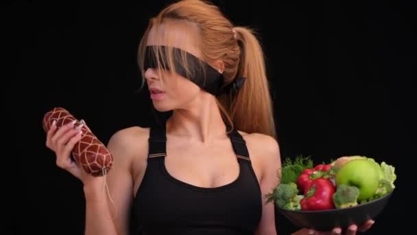 蒙着眼睛的女运动员一只手拿着熏肠 另一只手拿着蔬菜和蔬菜 — 图库视频影像
