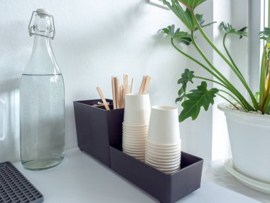 Cafe 'de self servis seti, bir şişe içme suyu ve beyaz arka plandaki beyaz tencerede yeşil yaprakların yanındaki kutuda beyaz kağıt bardak yığınıyla ahşap kahve çubuğu..