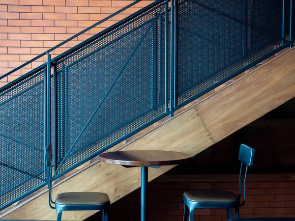 Cafe decoration design. Empty round wooden table with two modern bar stools near metal stairs on brick wall background.