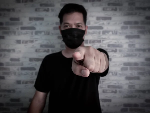 Asian serious man wearing black protective face mask and black shirt pointing finger at the camera on dark background, Coronavirus and pm 2.5 concept.