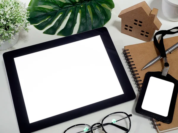 Work at home concept. White blank space screen on tablet, pen, notebook, glasses, green leaves, blank employee card and miniature wooden home on white background, top view.