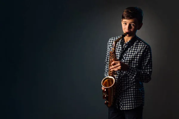 Un garçon sur un fond sombre avec un projecteur joue du saxophone — Photo