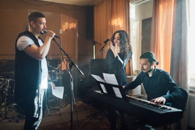 Elektronik klavyeye eşlik eden bir adam mikrofona şarkı söylüyor ve hoca da ses dersinde ağız şeklini gösteriyor.