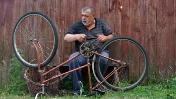 ひげを生やした年配の男性には木製のフェンスに座っているを修復する非常に古い自転車神経質 — ストック動画