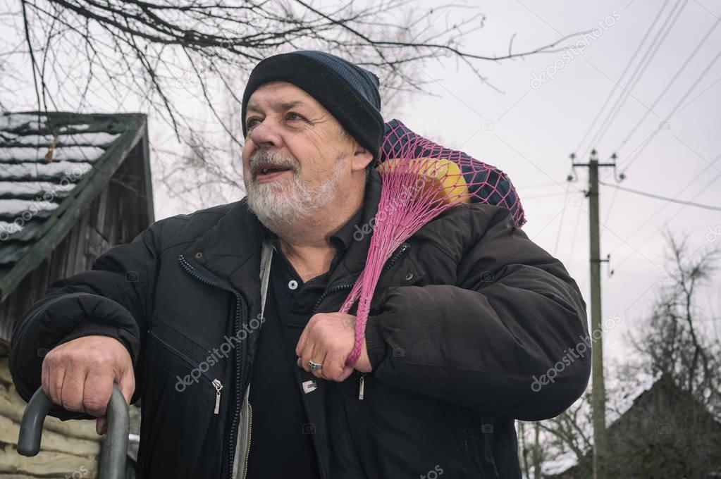 Nice portrait of positive elderly peasant with walking stick carrying some stuff in stringed bag while moving on an empty winter street of rural village in Ukraine