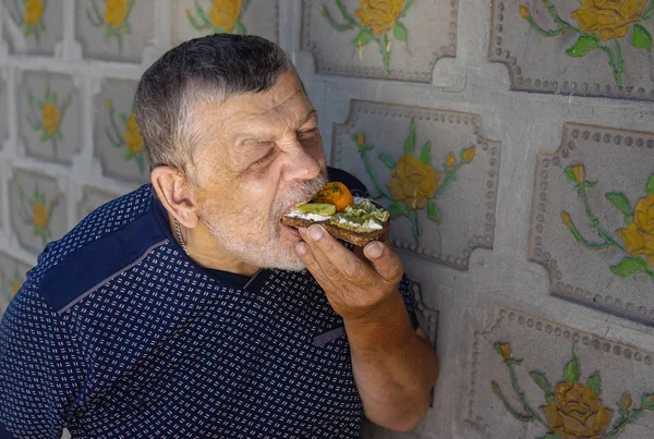 Portrait of hungry senior eating vegan sandwich with cucumber, yellow tomato and guacamole while standing against tiled wall