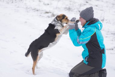 Mutlu olgun kadın kış mevsiminde karla kaplı bir zeminde siyah melez köpekle oynuyor.