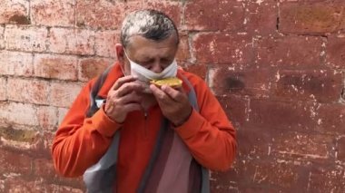 Gergin, Kafkas kıdemli bir adam kırmızı duvarın karşısında otururken sandviç yiyor. Covid 'de maske taktığını unutmuş. 19 salgın karantina dönemi. 