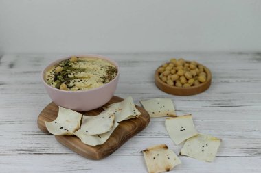 Humus, beyaz arka plan ve ahşap tahta üzerinde pembe bir tabakta geleneksel bir doğu mutfağıdır. Ev yapımı nohut, susam ezmesi, limon, tohum ve cips ile süslenmiş. Boşluğu kopyala.