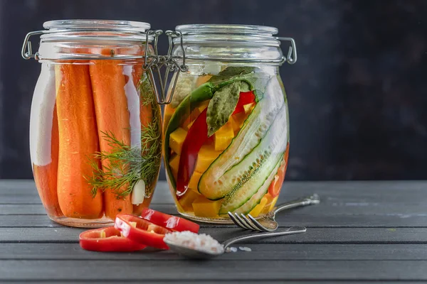 Две стеклянные банки ферментированных огурцов, морковь, перец, соль, овощи на темном фоне. популярная вегетарианская еда. копировальное пространство — стоковое фото