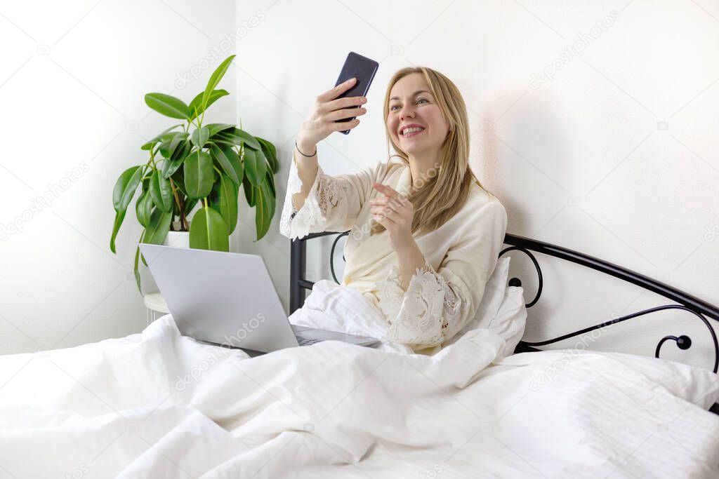 Young pretty european blonde woman working and studying from home, makes selfie and smiles, laughs . in bed with laptop and smartphone. she is in light, white, comfortable home clothes.