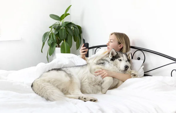 Açık renk elbiseli, sarı saçlı, gülümseyen genç bir kadın beyaz bir yatakta evcil bir hayvanla yatıyor. Sibirya Husky cinsinden köpeğiyle evde vakit geçiriyor. Tembel sabah