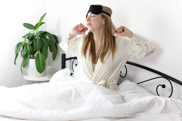 a European woman with blonde hair is sitting on a white bed, she just woke up. she is dressed in light clothing and a sleep mask. lazy weekend morning in a good mood