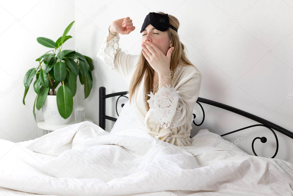 a European woman with blonde hair is sitting on a white bed, she just woke up in the morning. she is dressed in light clothing and a sleep mask. she did not sleep well and yawns