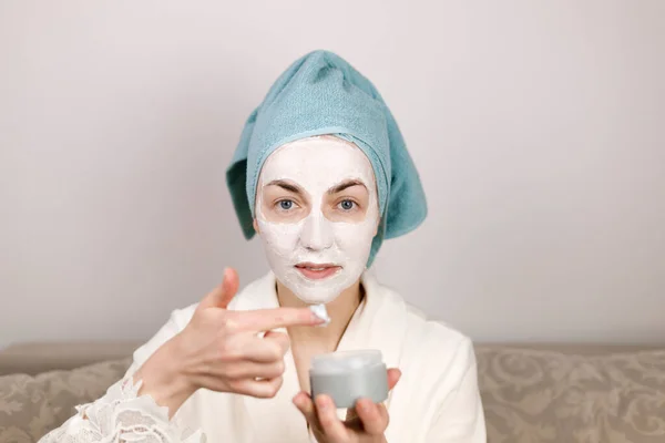 woman with a blue towel on her head applies a cosmetic mask of white clay to the face. mock up