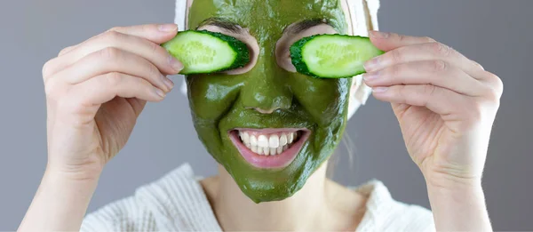Pankart. Kafasında havlu olan kadın salatalıklı yeşil kilden kozmetik bir maske yapıyor. Komik ev spa konsepti.
