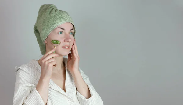 a European woman with a towel on her head makes self-massage of guasha with a jade roller. spa wellness concept. copy space