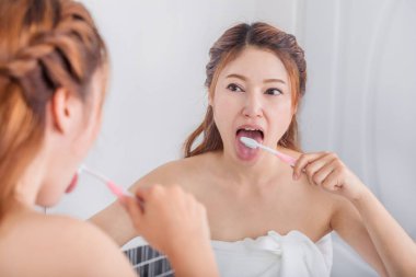 woman cleaning tongue using toothbrush with mirror in bathroom clipart
