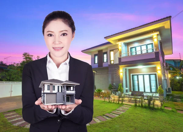 Model huis in zakelijke vrouw hand met de werkelijke huis en tuin — Stockfoto