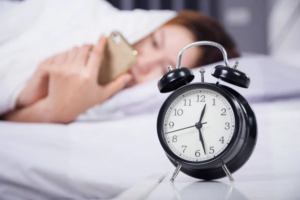 Clock with woman using her smartphone on bed — Stock Photo, Image