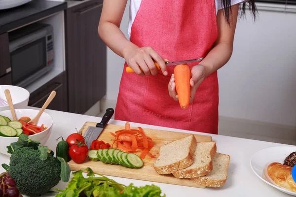 Mão de cenouras de casca de mulher com uma faca na sala de cozinha — Fotografia de Stock