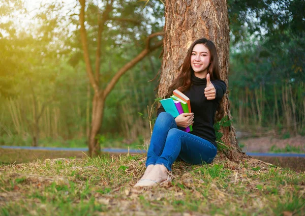 Giovane donna in possesso di un libro e mostrando pollice su segno nel parco — Foto Stock