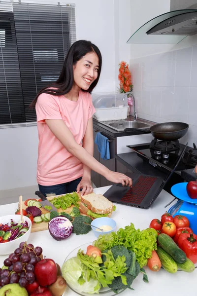 Mujer feliz cocina y mirando con el ordenador portátil en la sala de cocina —  Fotos de Stock