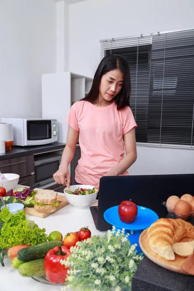 Donna che mescola insalata mentre cucina con il computer portatile in cucina — Foto Stock
