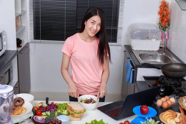Donna che mescola insalata mentre cucina con il computer portatile in cucina — Foto Stock