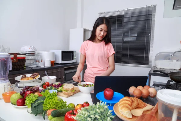 Donna che mescola insalata mentre cucina con il computer portatile in cucina — Foto Stock