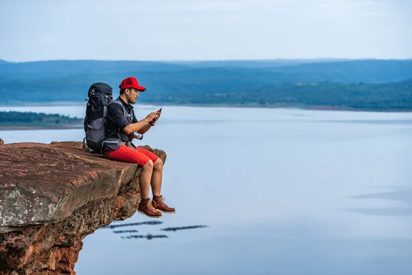 Hiker man sitting and using smartphone on edge of cliff, on a to — Stock Photo, Image