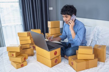 Genç adam akıllı telefondan konuşuyor ve evdeki bir yatakta online ürün satmak için dizüstü bilgisayar kullanıyor.