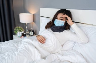 Tıbbi maskeli hasta kadın baş ağrısı ve virüsten muzdarip ve yatakta ateş, koronavirüs (covid-19) pandemik konsept.