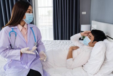 Doktor, yatakta hasta bir adamın sıcaklığını ölçmek için kızılötesi alın termometresi (termometre) kullanıyor, insanlar koronavirüsten (covid-19) korunan tıbbi maske takıyor olmalı.