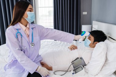 Doktor, yatakta hasta bir adamın sıcaklığını ölçmek için kızılötesi alın termometresi (termometre) kullanıyor, insanlar koronavirüsten (covid-19) korunan tıbbi maske takıyor olmalı.