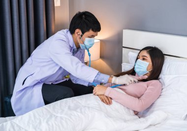Doktor, kadın hastanın kalbini dinlemek için steteskop kullanıyor. İnsanlar koronavirüsten (covid-19) korumak için tıbbi maske takıyor olmalı.