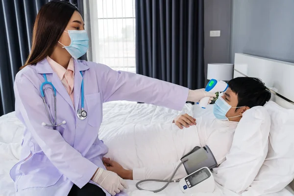 Doktor, yatakta hasta bir adamın sıcaklığını ölçmek için kızılötesi alın termometresi (termometre) kullanıyor, insanlar koronavirüsten (covid-19) korunan tıbbi maske takıyor olmalı.