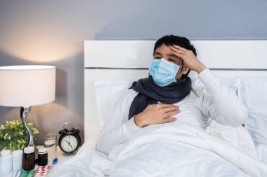 Tıbbi maskeli hasta adam baş ağrısı ve bir yatakta virüs hastalığı ve ateş, koronavirüs (covid-19) salgınından muzdariptir..
