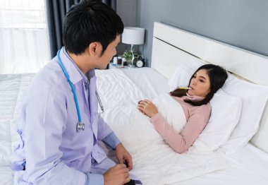 Doktor termometreyle ateşini ölçerken hasta kadın yatakta yatıyor.