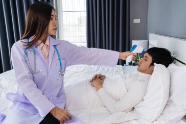 Doktor yatakta hasta bir adamın sıcaklığını ölçmek için kızılötesi alın termometresi (termometre tabancası) kullanıyor.
