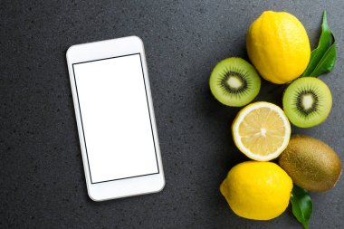 Limon ve kivi meyveli boş ekranlı akıllı telefon siyah taş masanın üstünde. Üst manzara, düz uzanma.