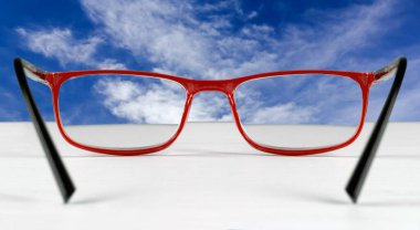 Mavi gökyüzü beyaz bir masada duran gözlüklerle görülüyor.