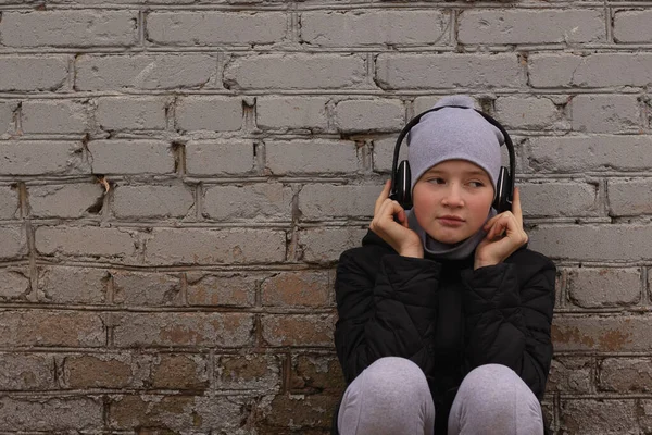 A teenager listens to music in black wireless headphones. Cool once and brick walls of houses. Modern children and technology